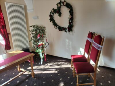 Im Trauzimmer mit großem Blumengesteck in Herzform an der Wand. Davor steht ein Kerzenständer mit Blumenschmuck und roten Kerzen. Rechts zwei Stühle für das Brautpaar.